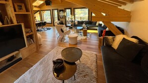 A spacious loft with high ceilings, wooden beams, a cozy seating area with a sofa and chairs, a flat-screen TV, and a dining area with a table and chairs.