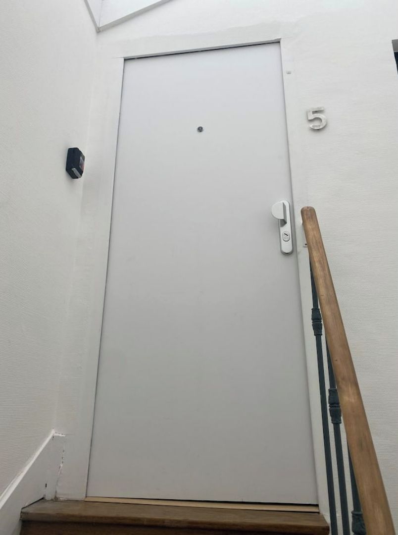 Een witte deur met een klein puntje in het midden, een deurbel, en een houten leuning aan de rechterkant.