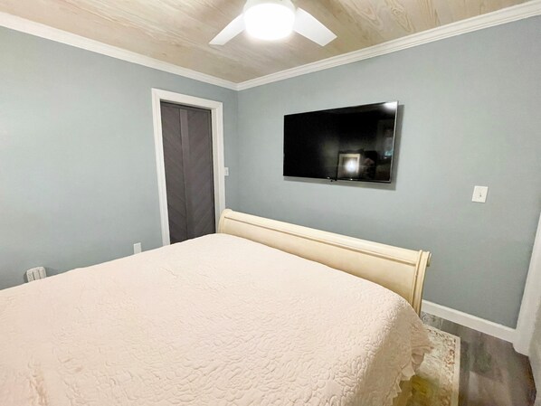 Un dormitorio con cama, ventilador de techo, televisor montado en la pared y una puerta.