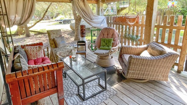 Uma área de estar coberta ao ar livre com móveis de vime, uma mesa de vidro e vista para uma rua arborizada.