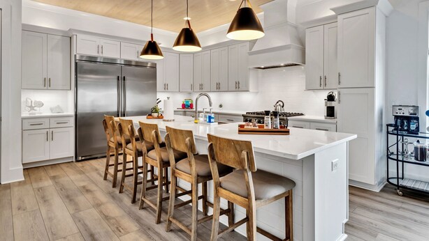 Una cocina moderna con armarios blancos, un refrigerador de acero inoxidable, una isla central con taburetes de bar, iluminación colgante y una máquina de café.