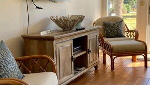 Een woonkamer met een aan de muur bevestigde televisie, een houten kastje met een decoratieve schaal erop, twee rieten fauteuils met kussens, en een groot raam met uitzicht op een gebouw en palmbomen.