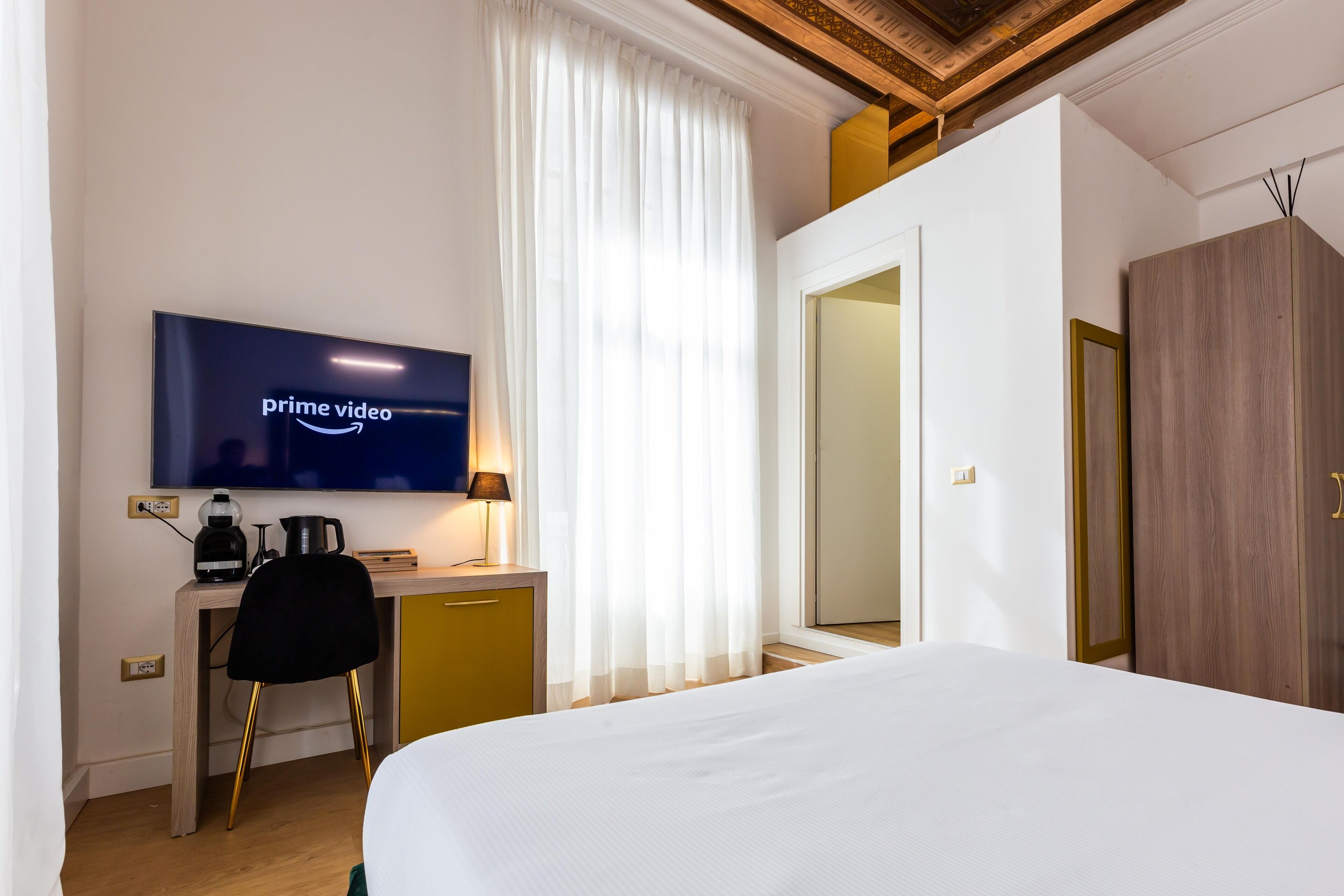A hotel room with a bed, a desk with a computer, a television displaying 'prime video', a coffee maker, and a wardrobe.