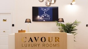 Un mostrador de recepción con la señalización "CAVOUR LUXURY ROOMS", un logotipo enmarcado arriba y una planta en maceta a un lado.
