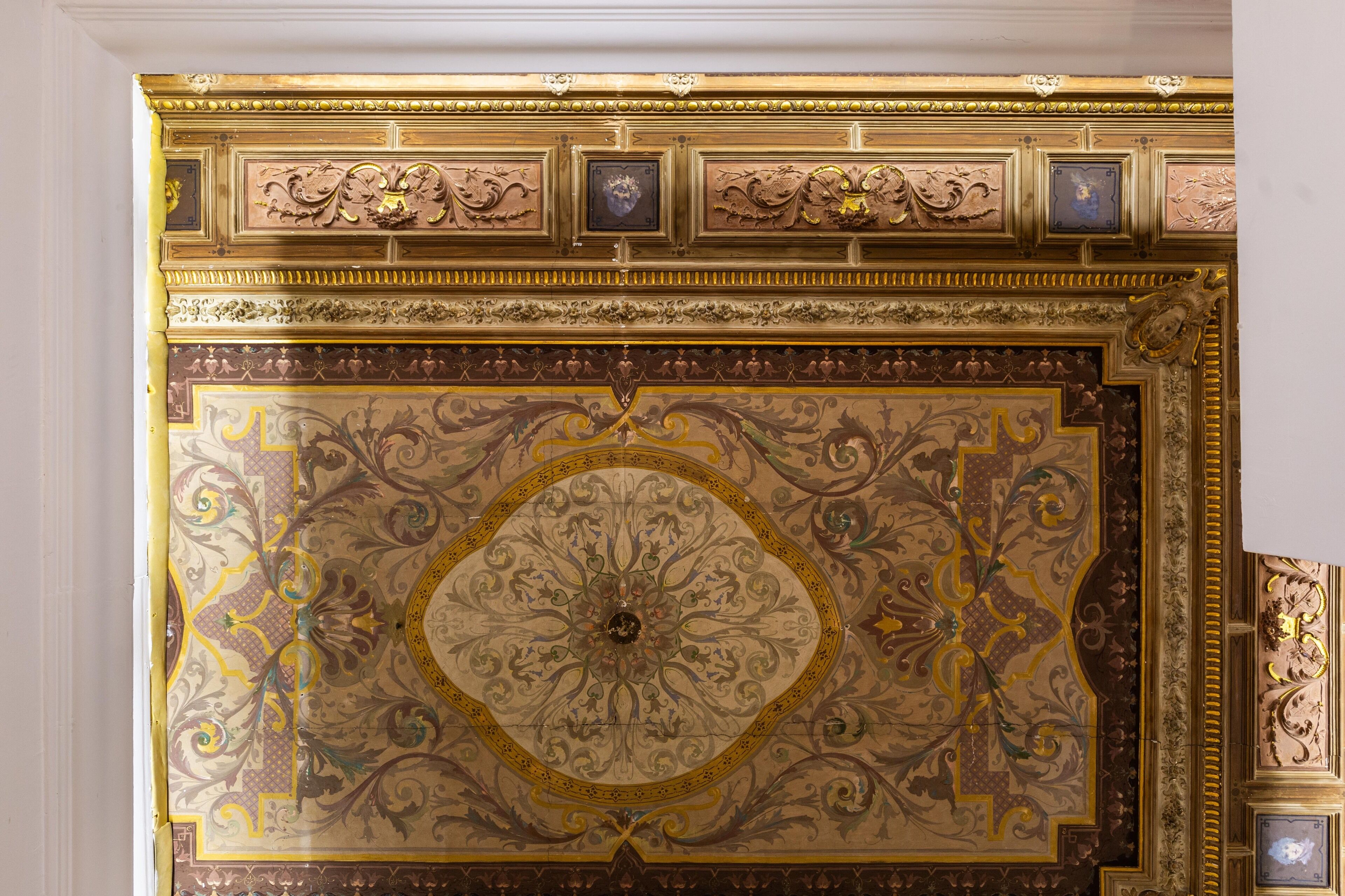Et detaljeret loft med udsmykkede guld- og brunmønstre, herunder en central rosette og indrammet kunst.