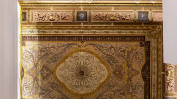 Et detaljeret loft med udsmykkede guld- og brunmønstre, herunder en central rosette og indrammet kunst.