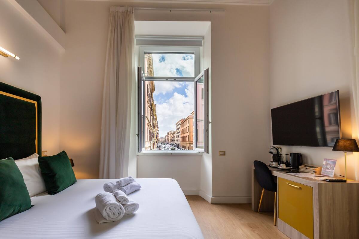 A hotel room with a bed, a desk, a chair, a TV, and a window with a city view.
