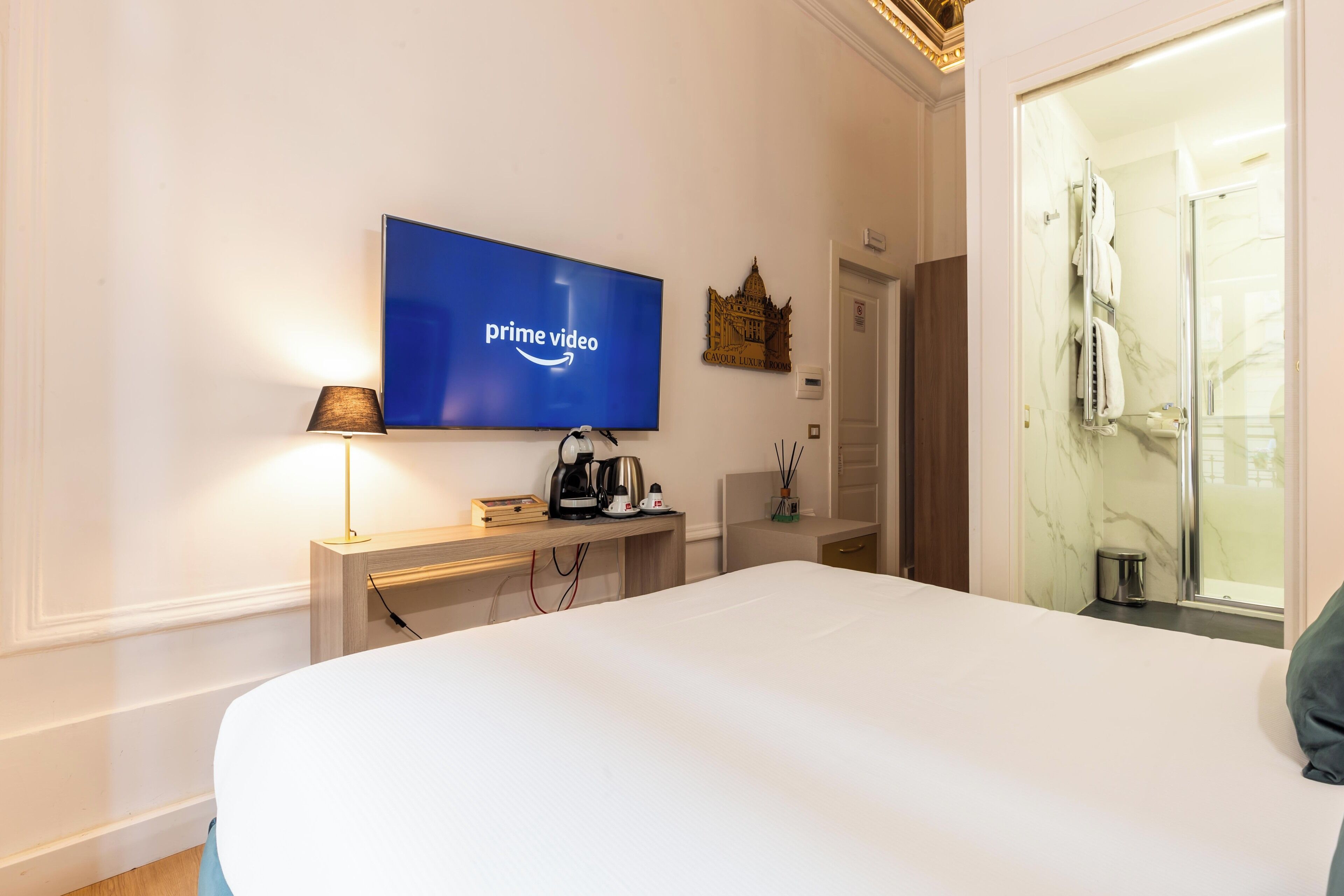 A hotel room with a bed, a television displaying the Prime Video logo, a bedside table with a lamp, a coffee maker, and a small decorative item.