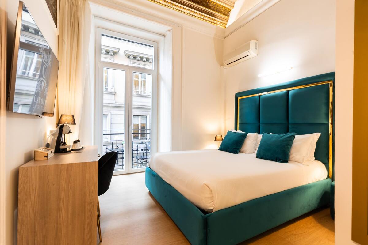 A modern hotel room with a large bed, a desk, and a view of the street through a glass door.