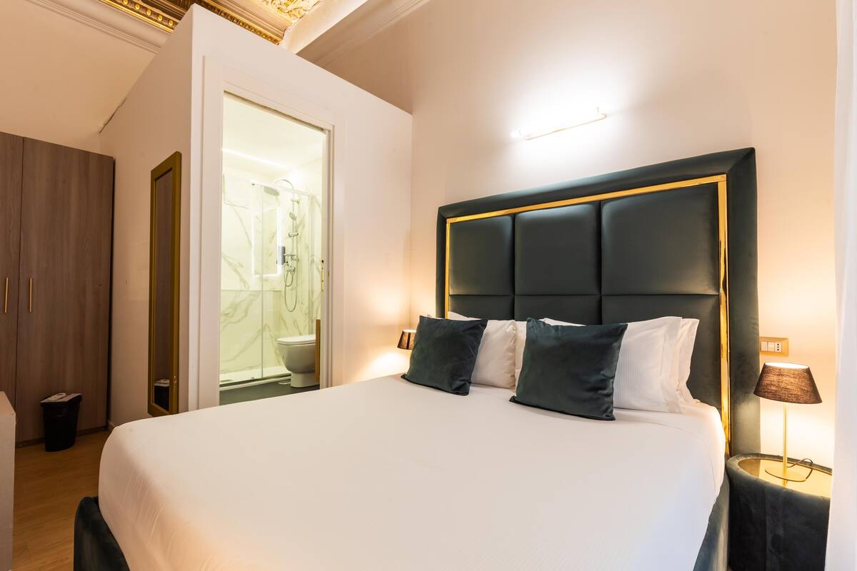 A hotel room with a large bed, a nightstand with a lamp, a wooden wardrobe, and a bathroom visible through an open door.