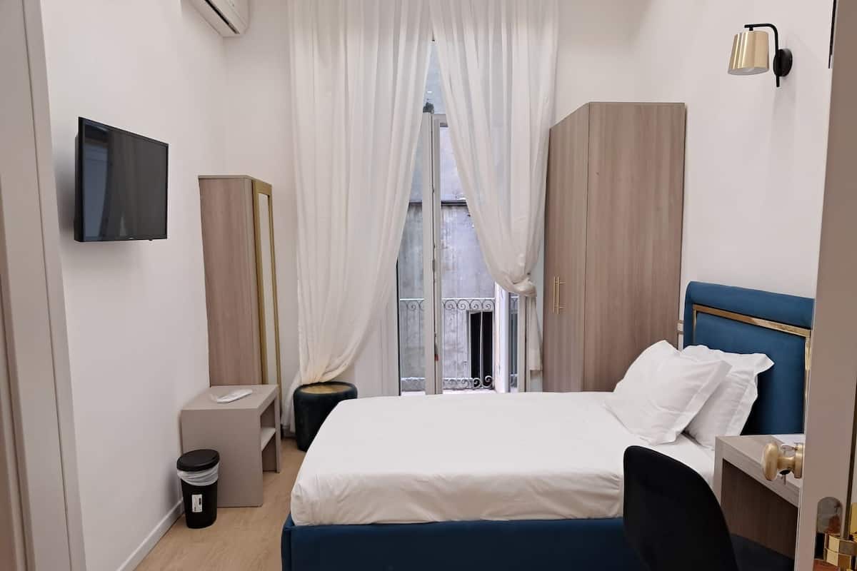A bedroom with a bed, a desk, a chair, a TV, a trash bin, and a window with curtains.