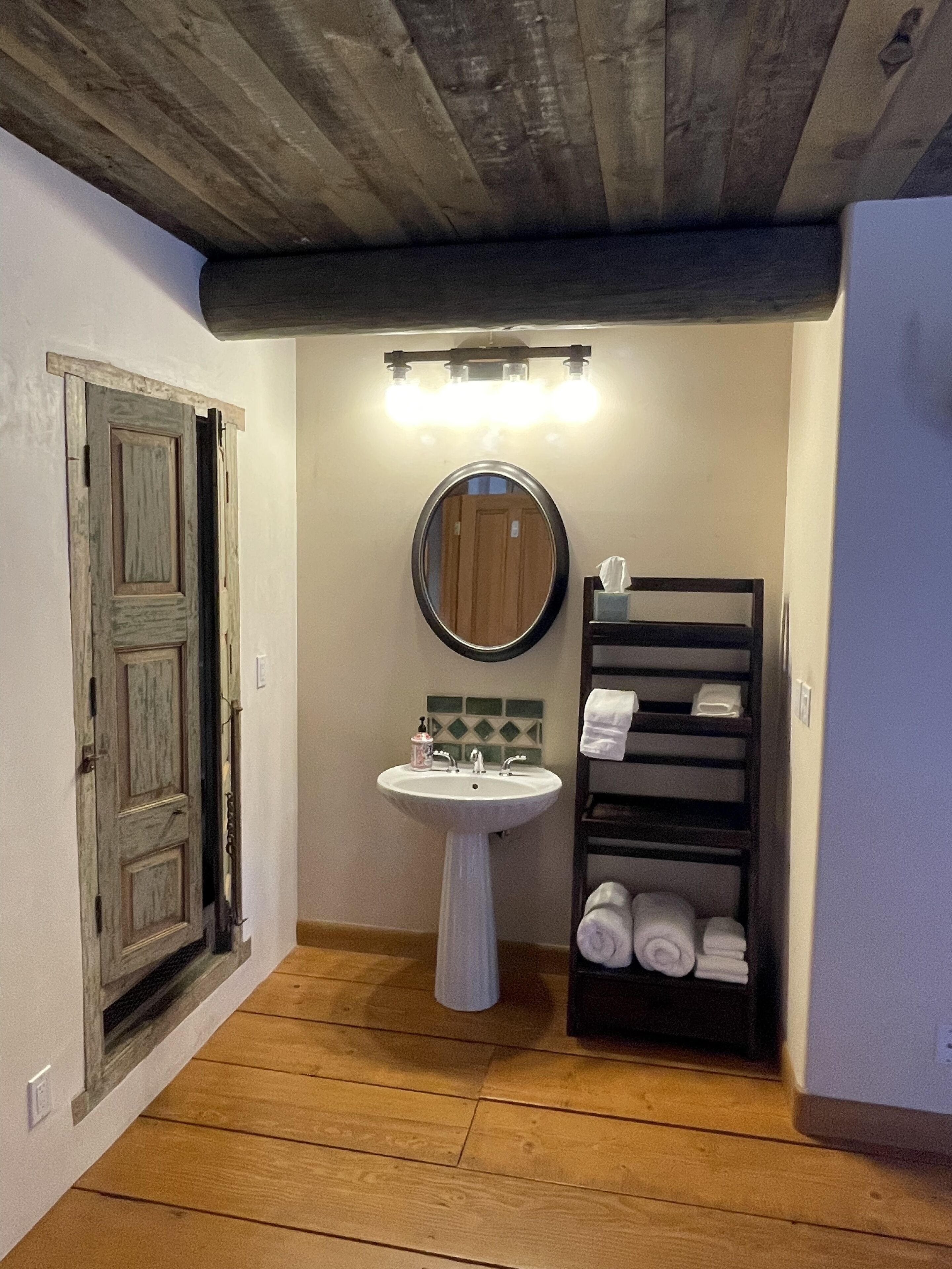 A bathroom with a wooden door, a pedestal sink, a mirror, and a towel rack with towels.