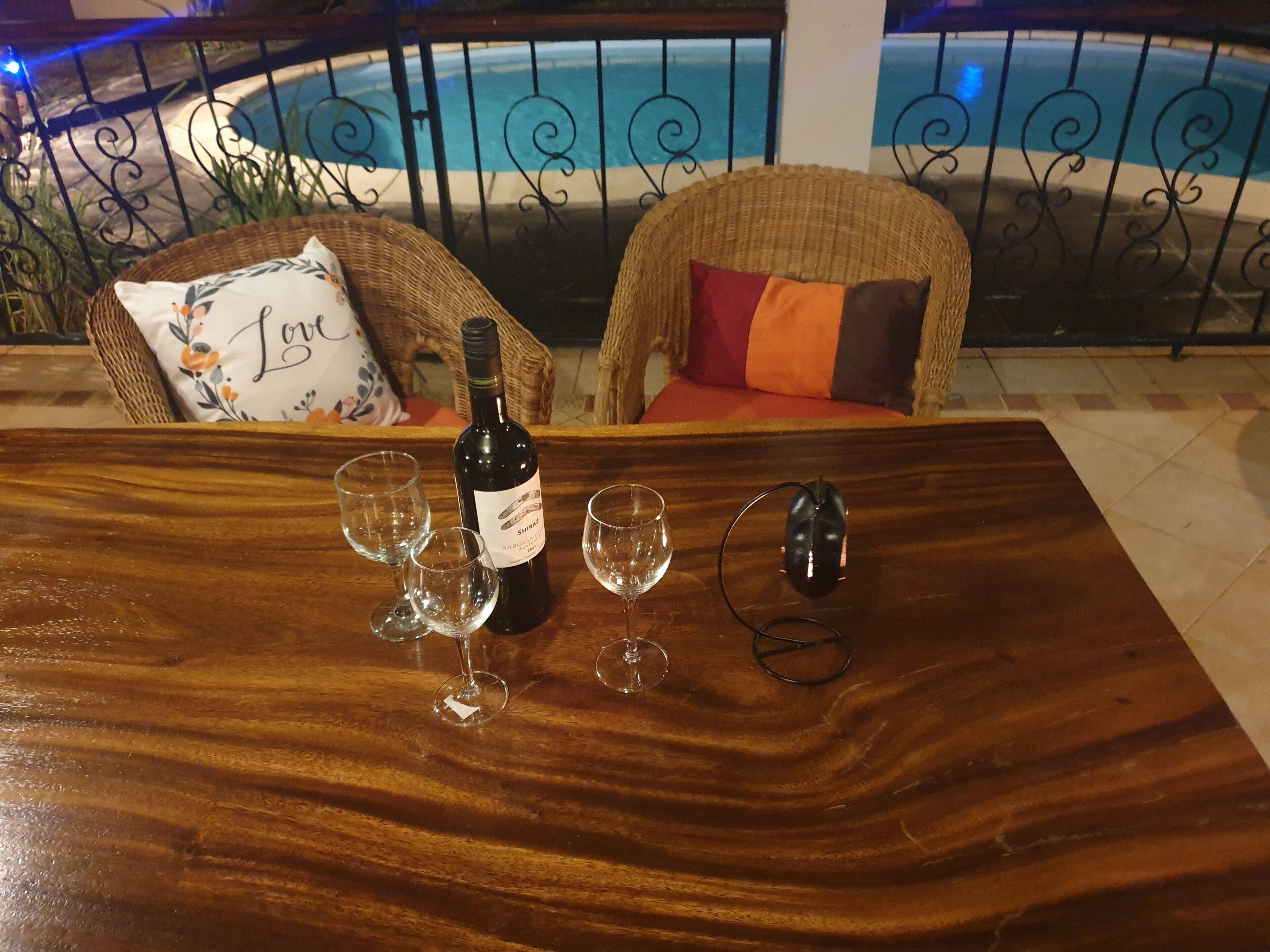 A wooden table with a bottle of wine, glasses, and a wine opener on a balcony with wicker chairs and a view of a swimming pool.