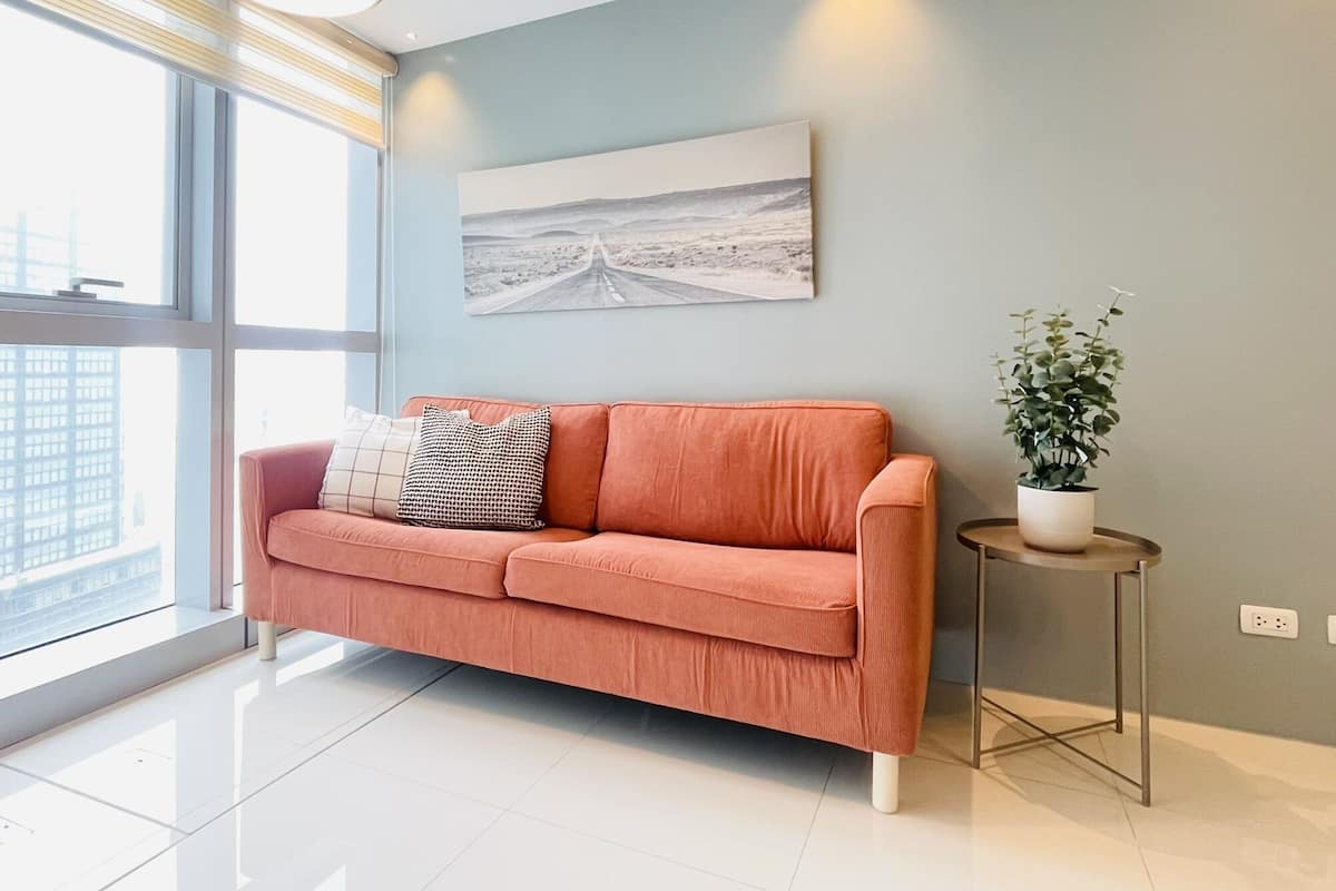 A modern living room with a sofa, a small side table with a plant, a painting on the wall, and a large window with blinds.