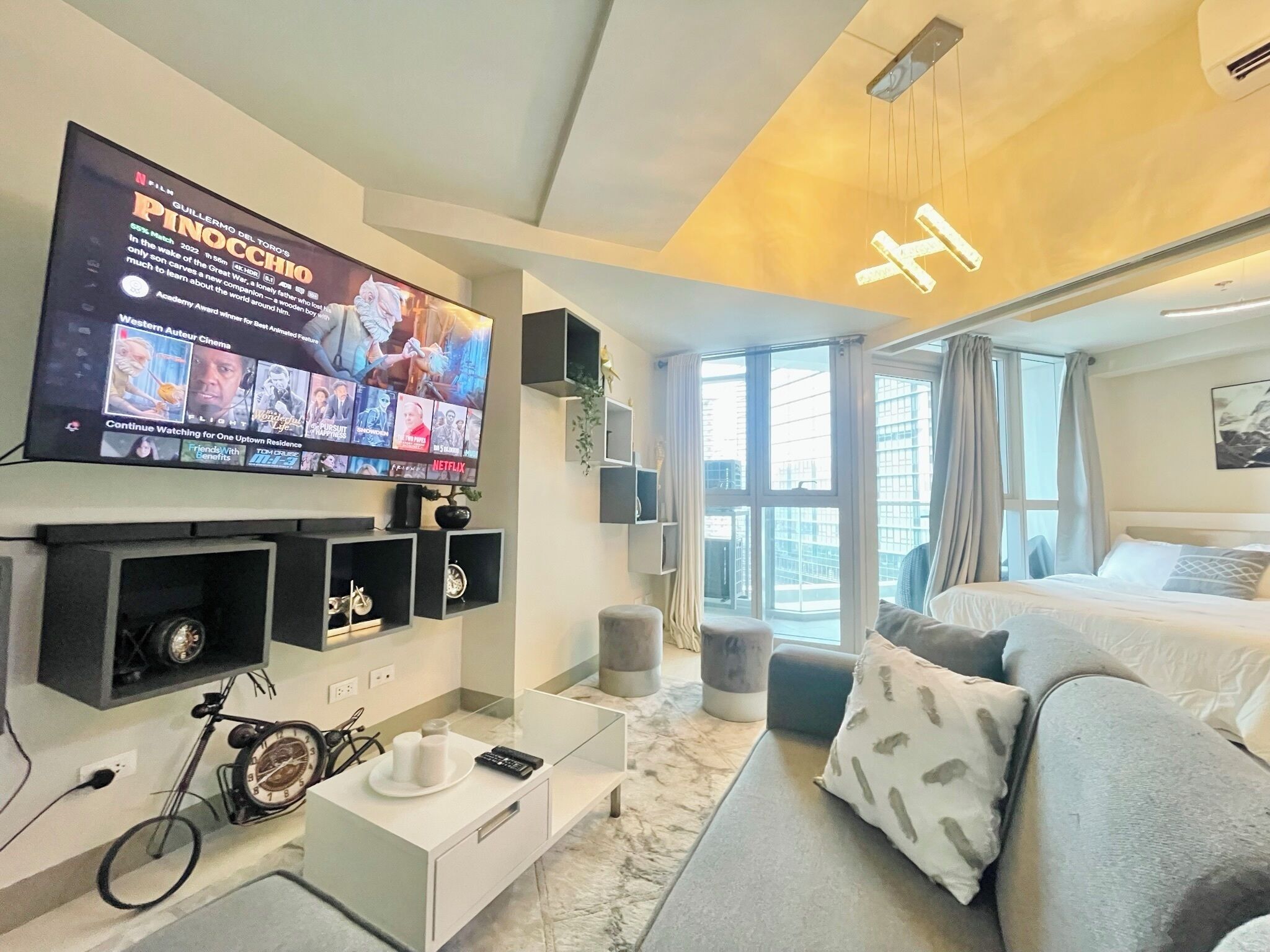 A modern hotel room with a large flat-screen TV, a sofa, a bed, and a window with a city view.