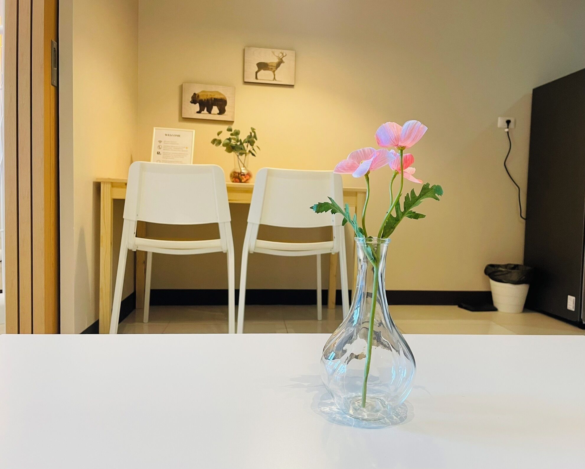 A room with a white table, two white chairs, a clear glass vase with flowers, and a welcome sign on the wall.