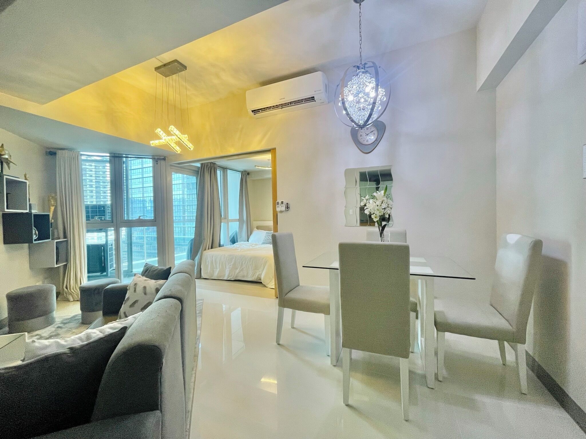 A modern hotel room with a bed, a dining table with chairs, a sofa, a TV, and a decorative light fixture.