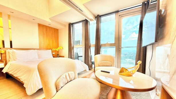 Ein modernes Hotelzimmer mit Bett, Sofa, rundem Couchtisch und großen Fenstern mit Blick auf die Stadt.