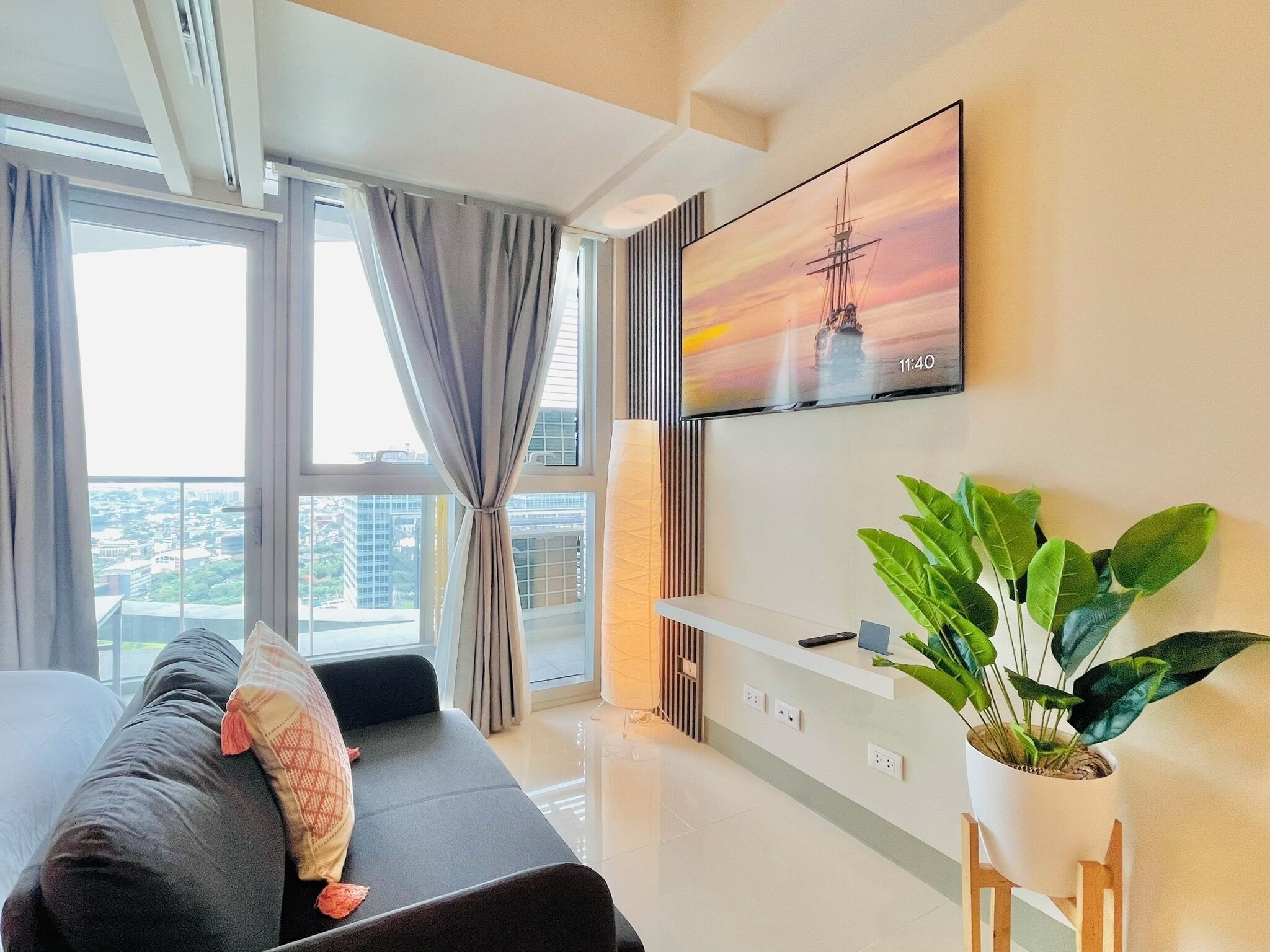 Una habitación de hotel moderna con un gran ventanal, un televisor de pantalla plana montado en la pared, un sofá con cojines y una planta en maceta.
