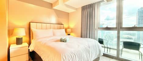 A modern hotel room with a large bed, two bedside lamps, a view of the city through large windows, and a balcony with chairs.