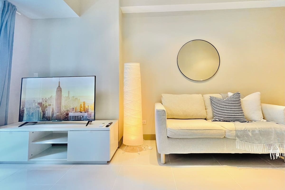 A modern living room with a flat-screen TV, a white sofa, a round mirror, and a tall white lamp.