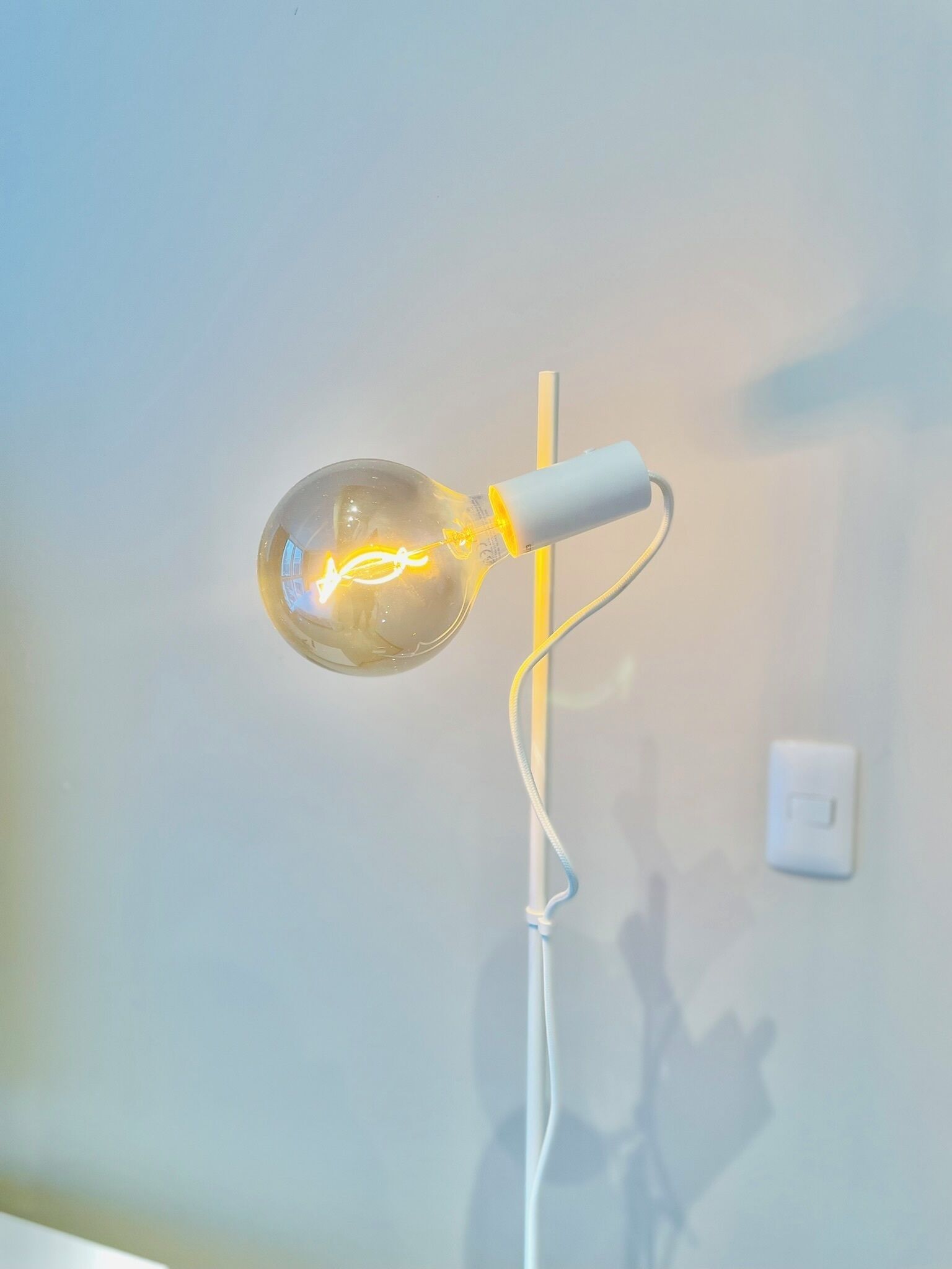 A single light bulb with a visible filament, connected to a white power cord, mounted on a wall.
