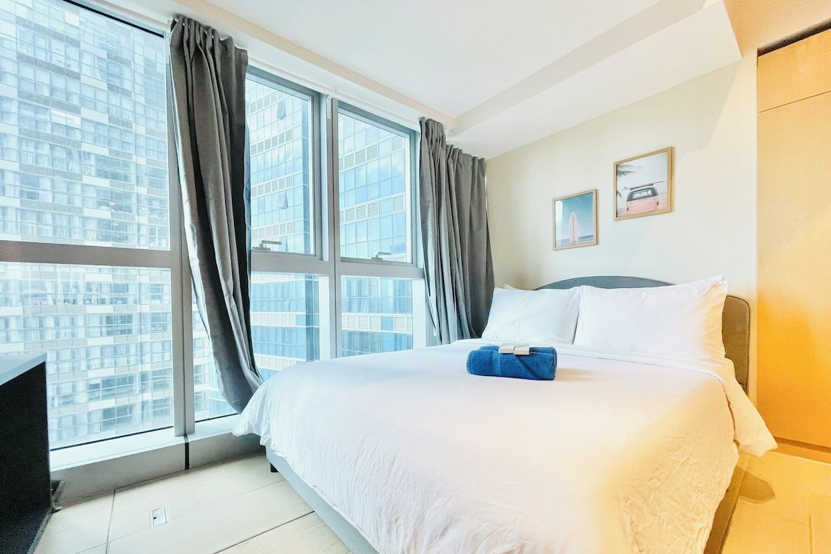 A hotel room with a large bed, a desk, and a view of a cityscape through large windows.