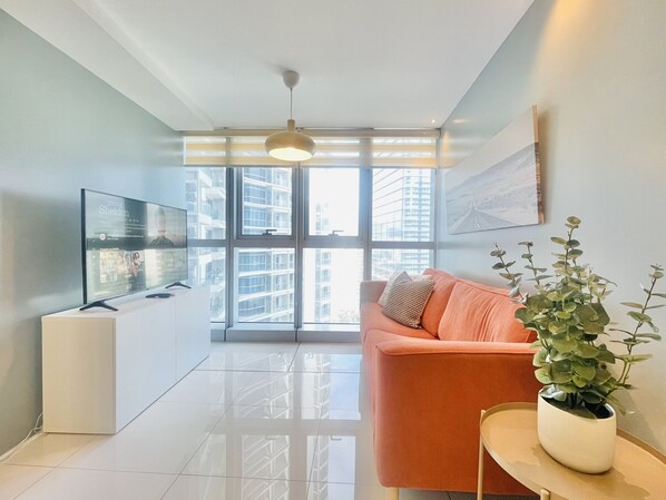 A modern living room with a sofa, a TV, a plant, and a view of a cityscape through large windows. - Uptown Parksuites 1 (Taguig)