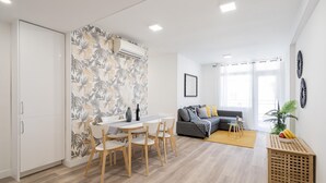 A modern living room with a sofa, a dining area with a table and chairs, and a kitchen area with a white cabinet and a patterned wall.