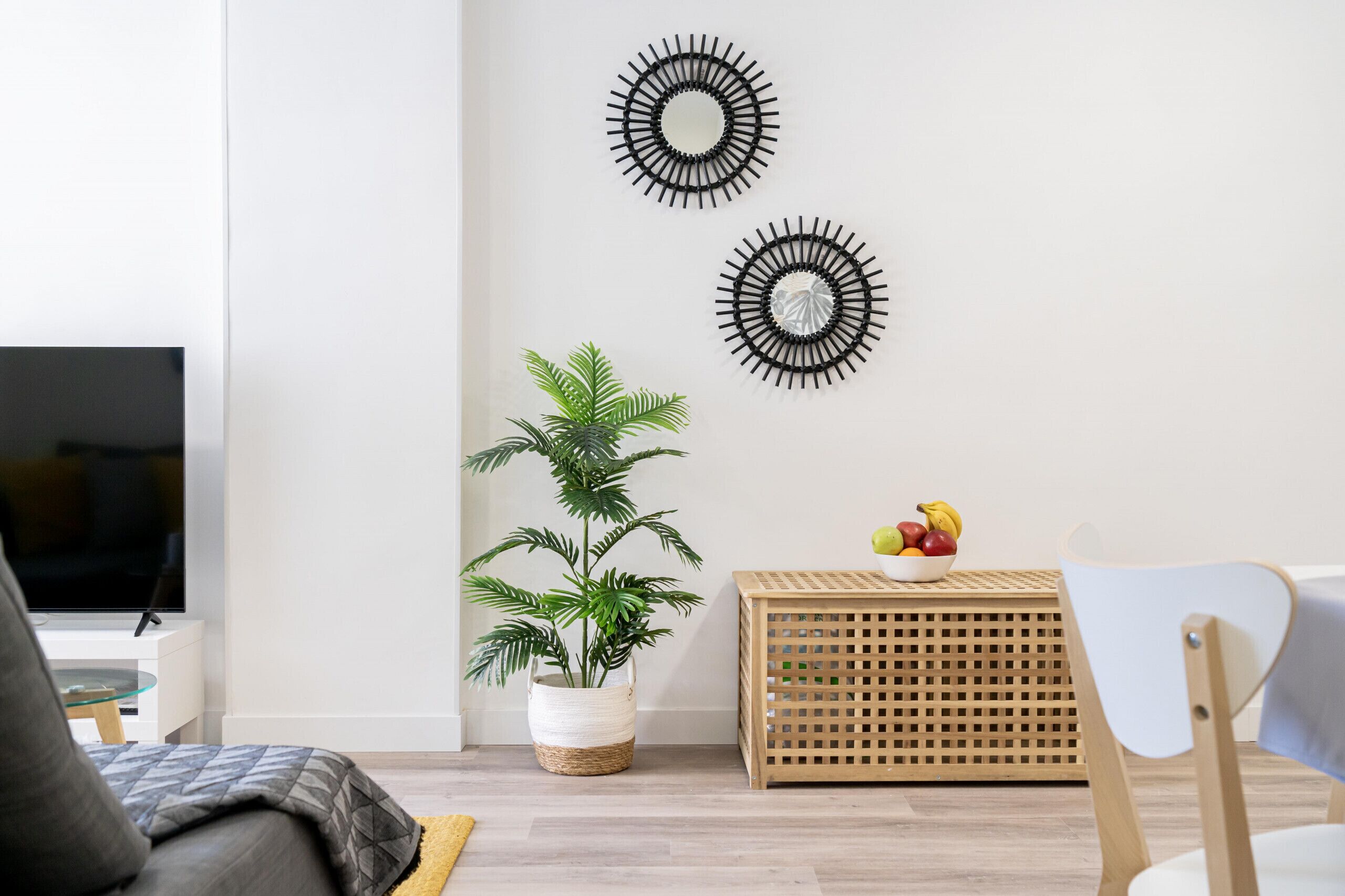 Un salon moderne doté d’un téléviseur à écran plat, d’une plante en pot, de deux miroirs décoratifs fixés au mur et d’une table d’appoint en bois sur laquelle sont disposés des fruits.