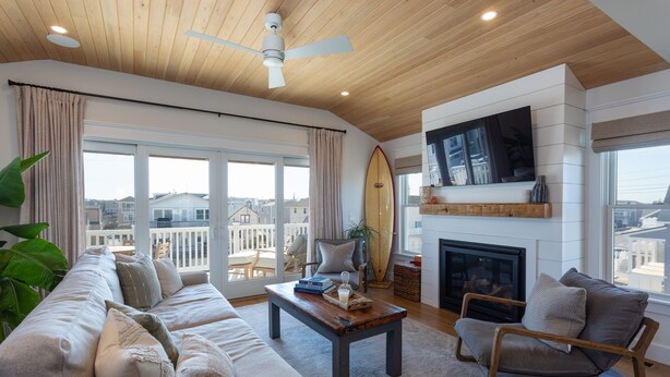 A modern living room with a wooden ceiling, a large window with a view, a fireplace, and a surfboard on the wall. - Oceanside home - 2 min walk to the beach! Ocean & bay views! (Ship Bottom)