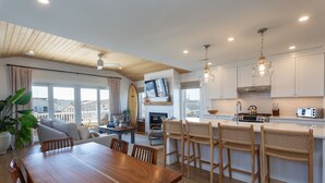 A modern kitchen with wooden cabinets, a dining area with a wooden table and chairs, and a large window with a view of buildings. - Oceanside home - 2 min walk to the beach! Ocean & bay views! (Ship Bottom)