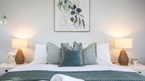 Ein ordentlich bezogenes Bett mit einer grünen Tagesdecke, zwei Nachttischlampen und einem gerahmten Pflanzenbild über dem Bett.