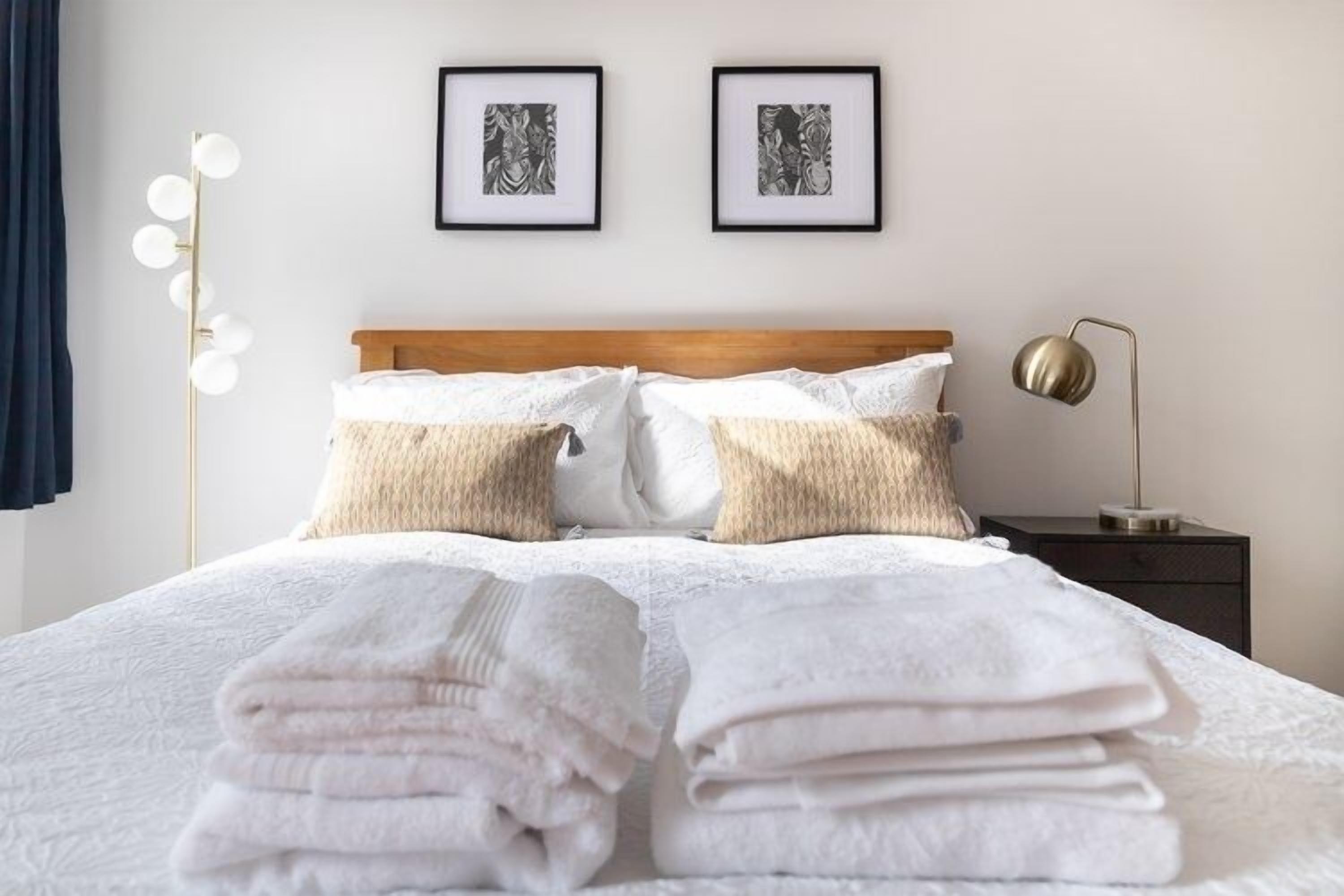 Ein ordentlich bezogenes Bett mit weißen Leinen und zwei Kissen, ein Nachttisch mit Lampe und zwei gerahmte Bilder an der Wand.