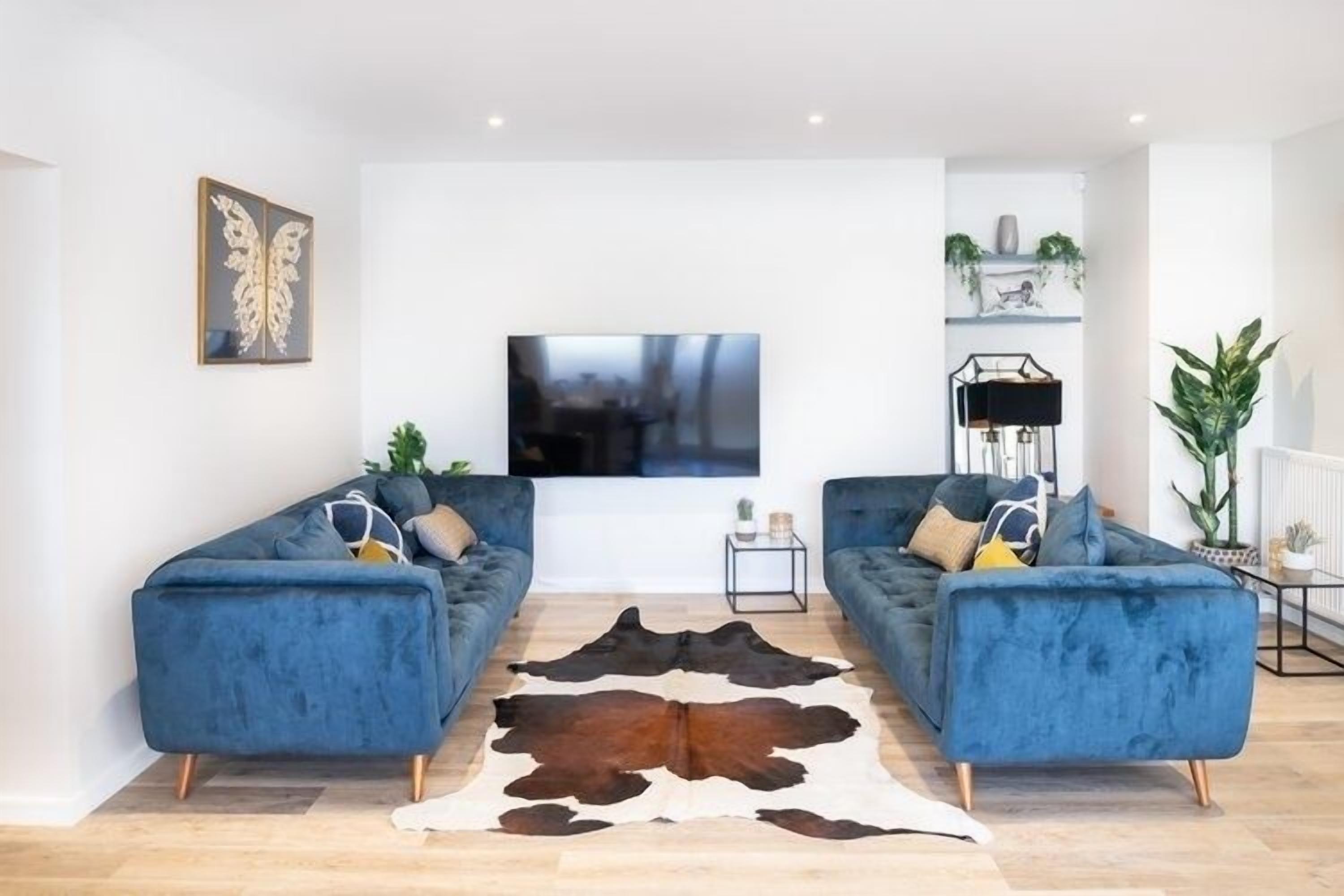 En moderne stue med to blå sofaer, et stort fladskærms-tv, en kohudes måtte og minimalistisk indretning.