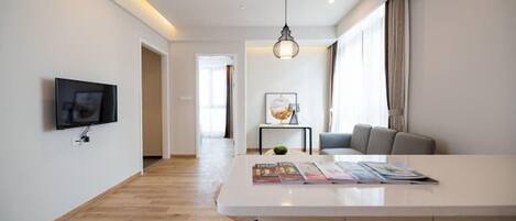 A modern living room with a flat-screen TV, a white coffee table with magazines, a hanging light fixture, and a sofa.
