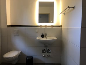 Uma casa de banho moderna com uma sanita, uma pia branca e um espelho com luz em cima.