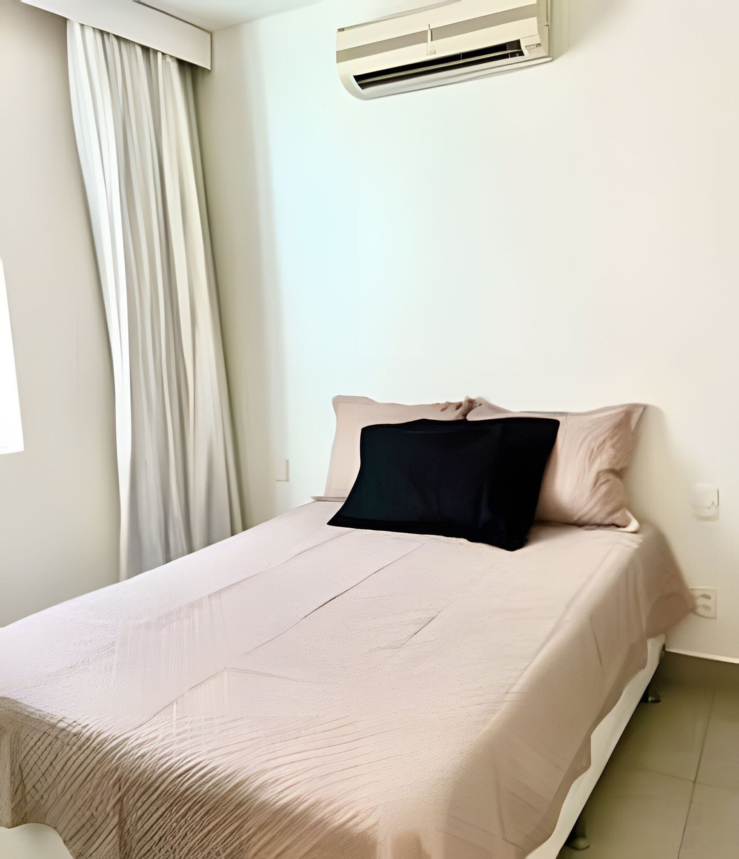 A single bed with a beige bedspread and a black pillow in a room with a white wall and an air conditioning unit above the bed.