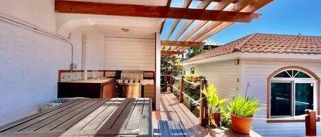 A covered patio area with a wooden table and chairs, a grill, and a small house with a tiled roof.