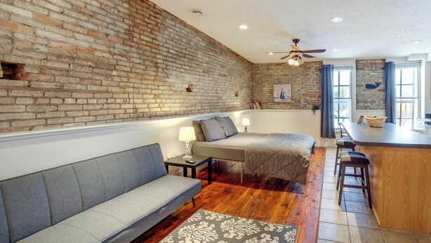 Una habitación acogedora con pared de ladrillo, un sofá, una cama, una mesita de noche con lámpara y una zona de bar con taburetes.