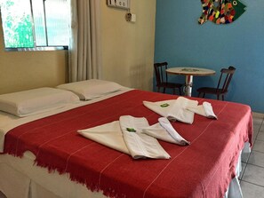 Habitación de hotel con una cama, una manta roja y toallas sobre la cama. En la esquina, hay una mesita redonda y dos sillas.