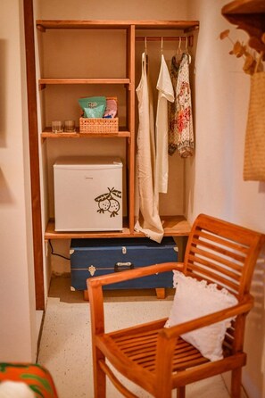 Una habitación compacta con un armario de madera, un refrigerador blanco pequeño, un contenedor azul y una silla de madera.