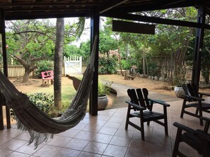 A covered patio with a hammock, wooden chairs, and a view of a garden with a wooden fence and trees.