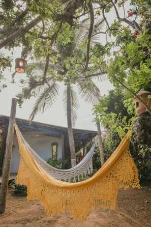 A hammock suspended between trees in a garden area with a building in the background. - Ecocharme Pousada Netuno (Fernando de Noronha)