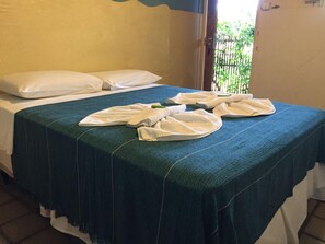 Habitación de hotel con cama, almohadas blancas, colcha color turquesa y ventana con vista a vegetación.