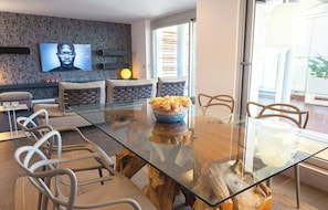 Una sala moderna con una mesa de comedor de vidrio, asientos cómodos, un televisor montado en la pared y vistas a través de la puerta de cristal.
