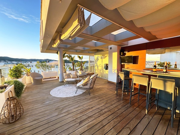 A wooden deck with outdoor seating, a bar area, and a view of the cityscape.