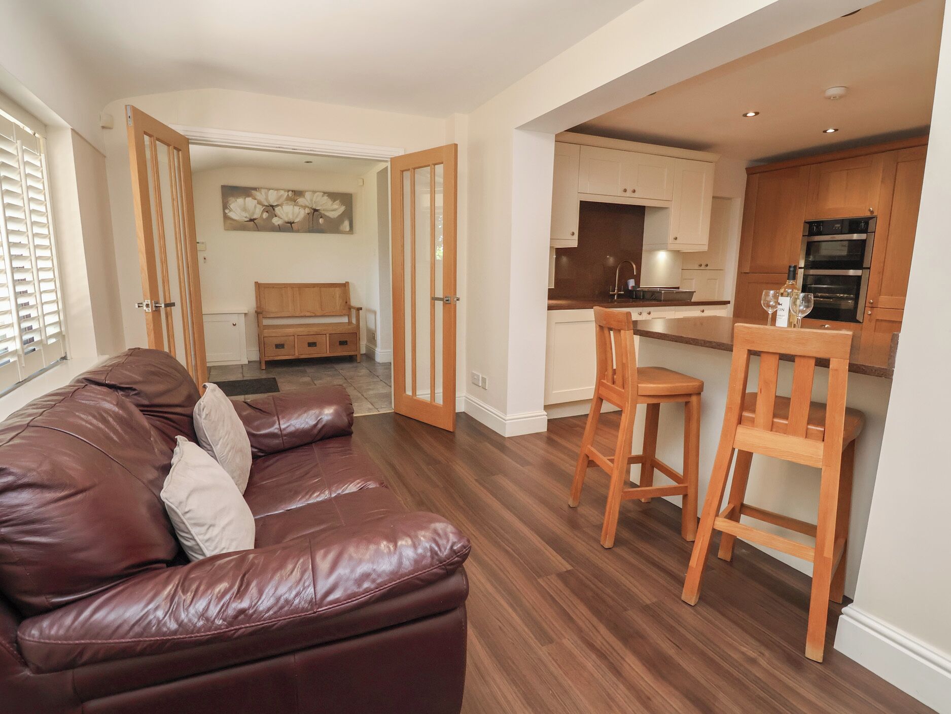 A modern living room with a leather sofa, a wooden bench, and a kitchen area with wooden cabinets and a bar stool.