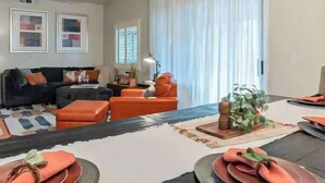 A modern living room with a sofa, orange chairs, a dining table set for four, and a ceiling fan.