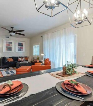 Uma sala de estar moderna com sofá, poltronas laranjas, uma mesa de jantar posta para quatro pessoas e um ventilador de teto.